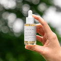 Hand holding a bottle of Veluma skincare serum with a blurred green background